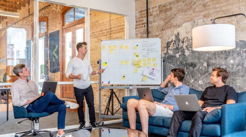 three men sitting while using laptops and watching man beside whiteboard